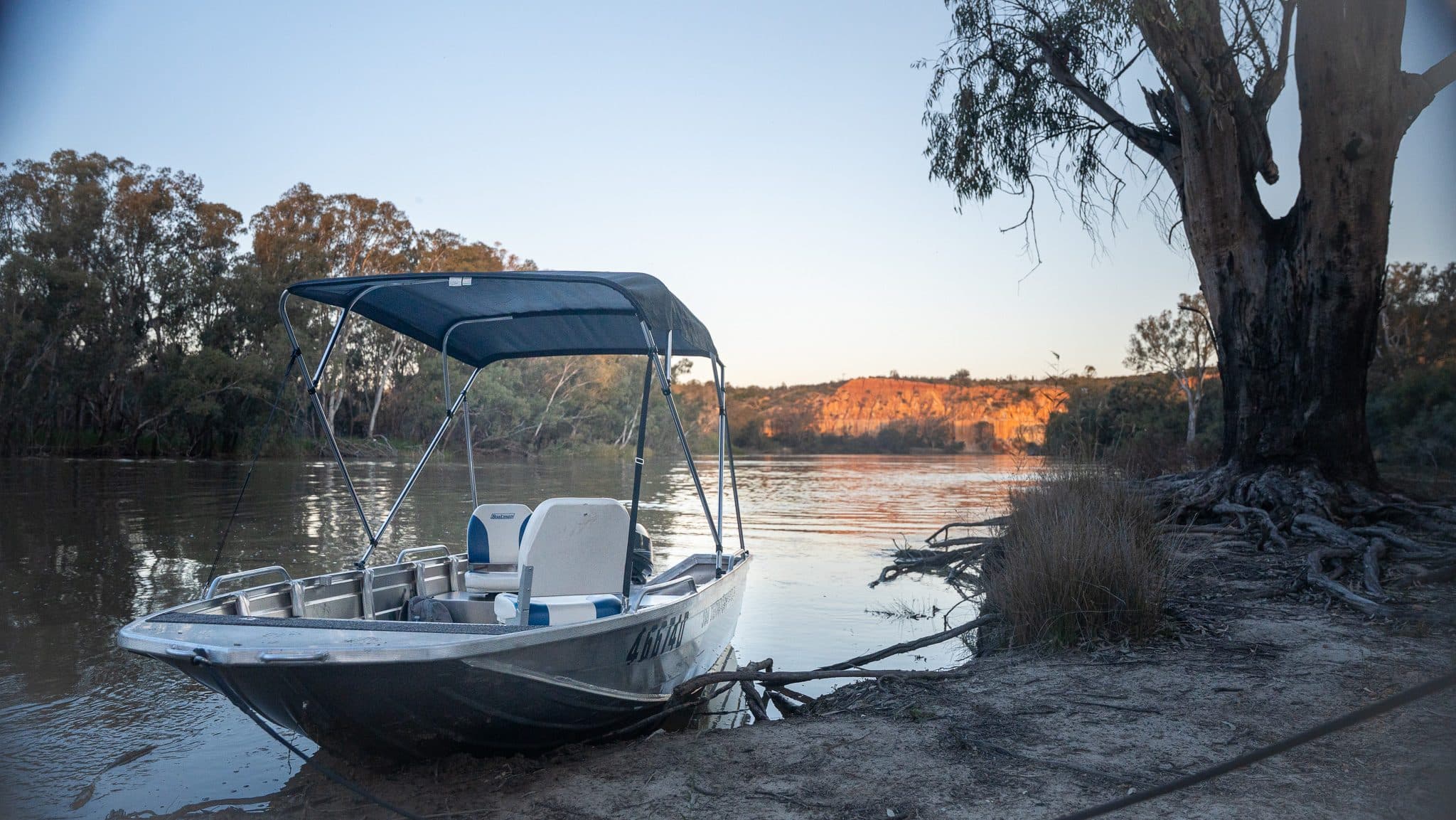 A silver Stacer Territory hire tinny with its bimini up, nosed onto the riverbank beneath a red gum, with ochre cliffs across the water