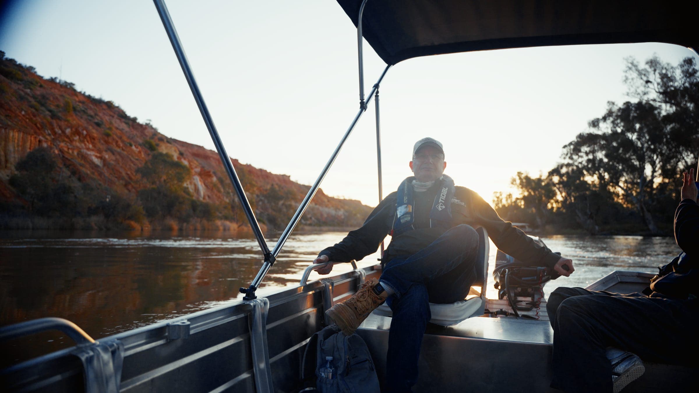 A man in a cap and lifejacket at the tiller of a silver hire tinny under its bimini, with red cliffs on one bank and river red gums against a low sun on the other