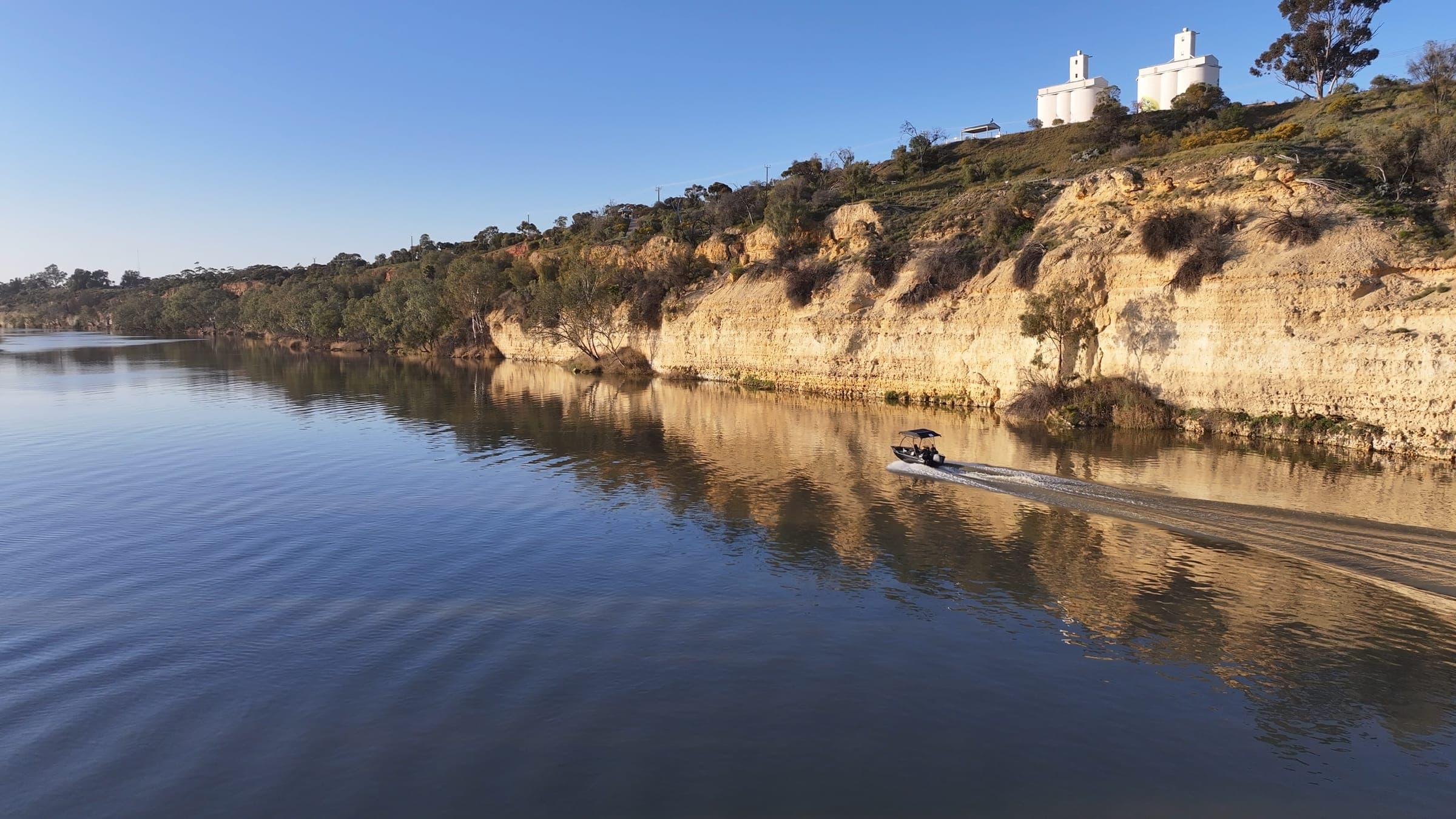 A tinny cruising beneath sunlit limestone cliffs, with grain silos on the clifftop