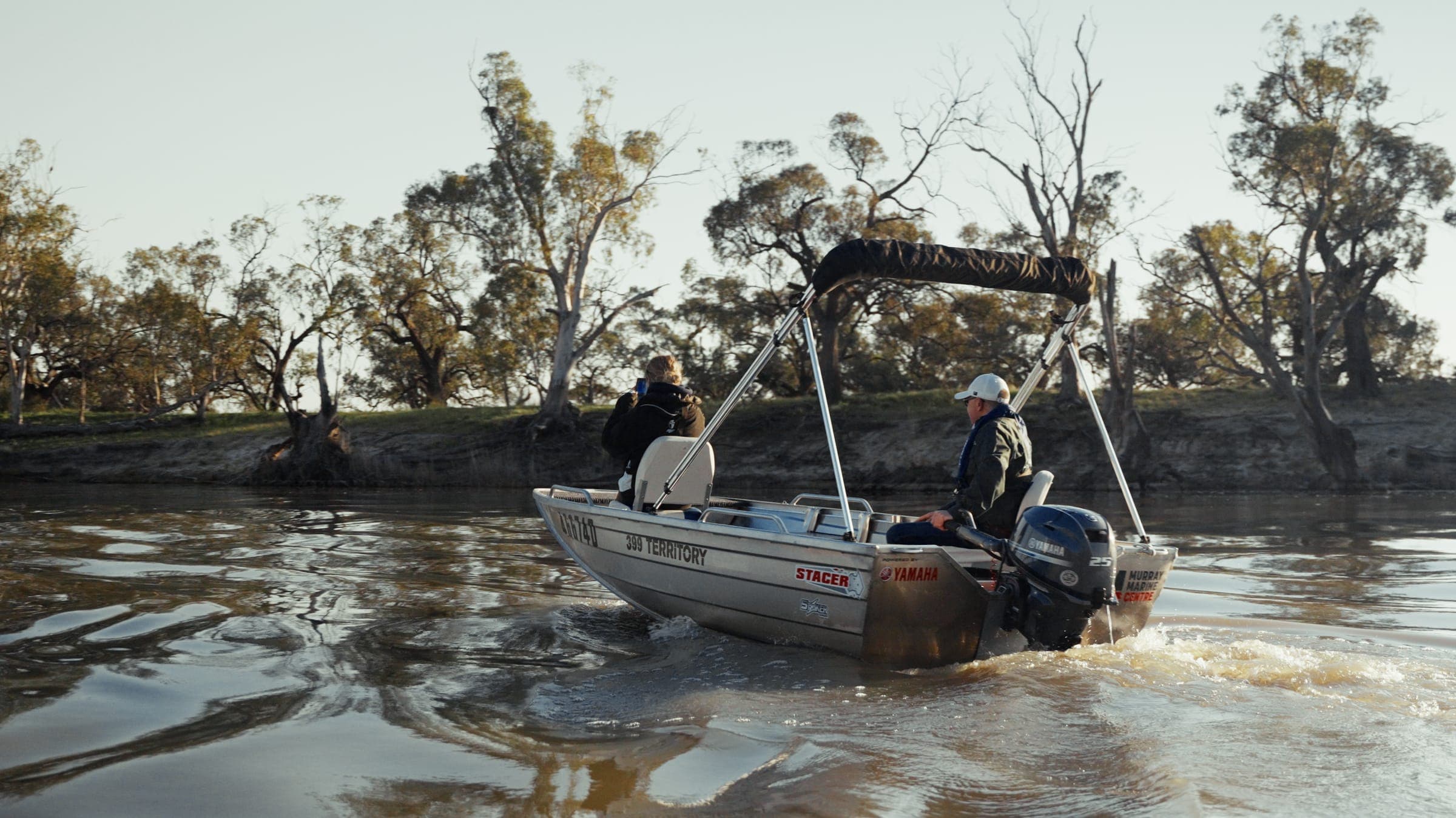 A hire tinny heading along the Murray River past river red gums, one passenger taking a photo